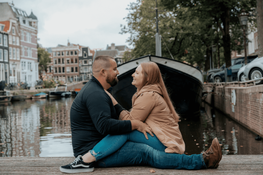 could sitting on the ground in front of an Amsterdam Canal. their legs are crossed over each other and they are smiling